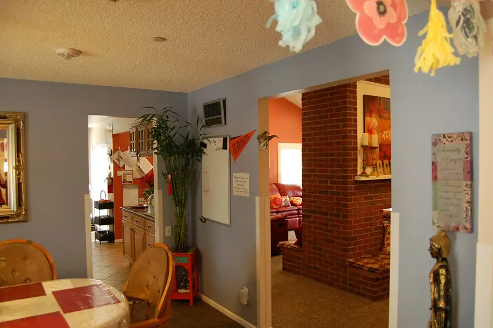Interior view of a house showing a dining area, kitchen, and living room with brick wall, blue walls, and various decorations including hanging flowers and a whiteboard.