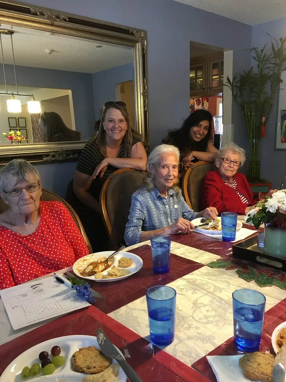 Four older women sit at a dining table with food and drinks, while two younger women stand behind them, all posing for a photo in a home dining room.