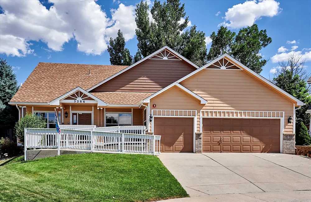 Single-story tan house with a three-car garage, white railing, front porch, sloped roof, and a green lawn under a partly cloudy sky.
