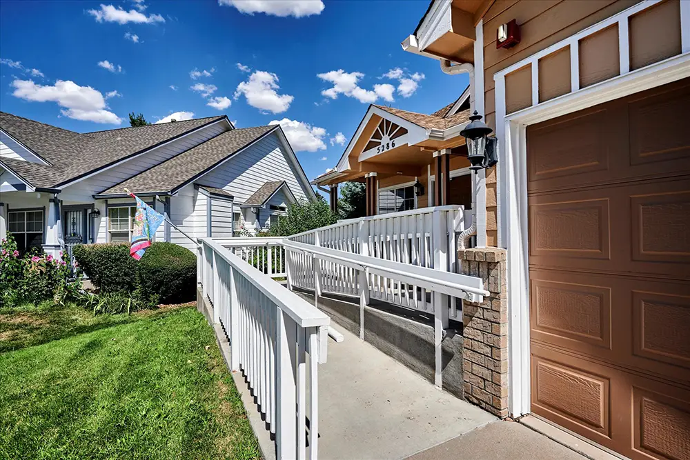 A concrete wheelchair ramp with white railings leads to the front entrance of a house with a brown garage door on a sunny day.