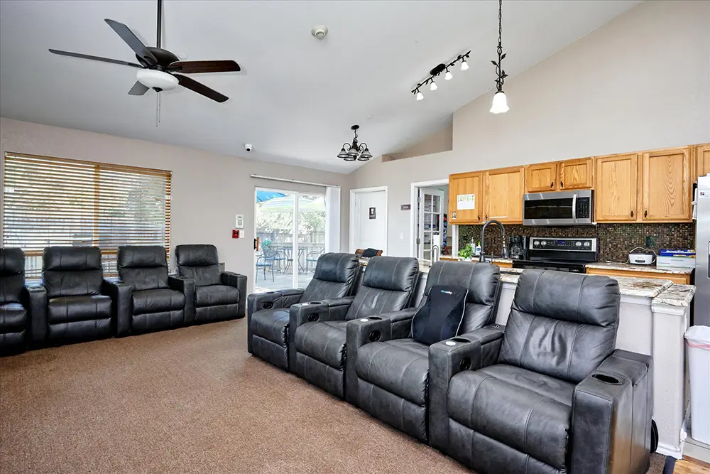 Room with eight black leather recliners facing a TV, brown carpet, ceiling fans, and an open kitchen with wooden cabinets and modern appliances.
