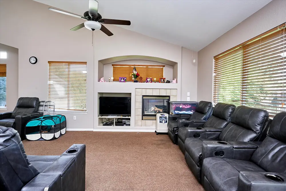 Living room with black recliner sofas, ceiling fan, TV, fireplace, brown carpet, two pet playpens, and large windows with wood blinds.