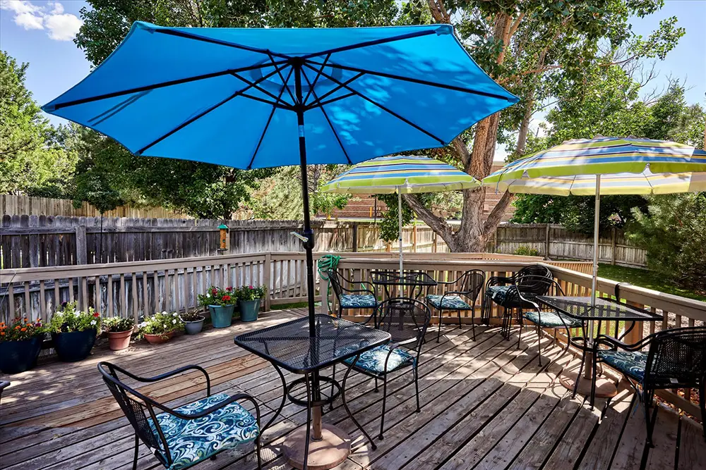 Outdoor wooden deck with potted plants, four metal tables and chairs, and three large patio umbrellas providing shade.