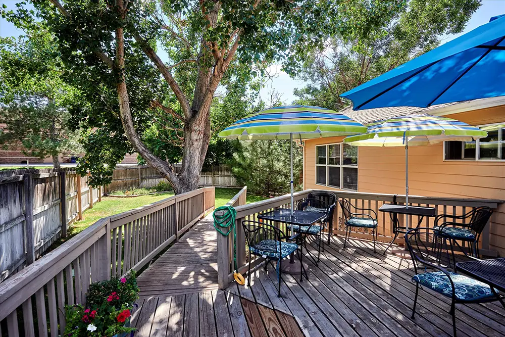 Wooden backyard deck with patio tables, chairs, and umbrellas, surrounded by trees and a wooden fence next to a yellow house.