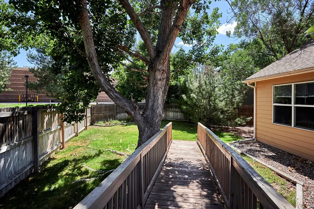 Wooden ramp with railings leads from a house to a grassy backyard with a large tree and fenced perimeter under a clear blue sky.