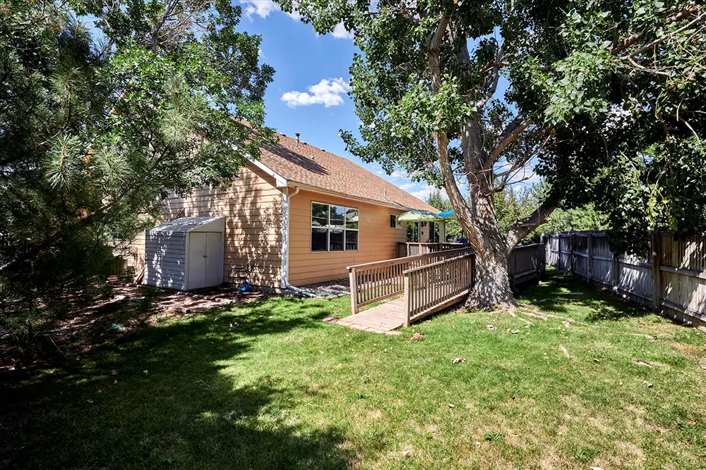 Single-story tan house with a sloped roof, backyard lawn, large shade tree, wooden ramp, shed, and wooden fence under a blue sky.