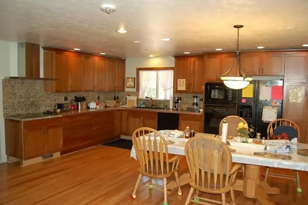 Spacious kitchen with wooden cabinets, granite countertops, and a dining table with four chairs. Appliances and utensils are visible on counters and table. Bright lighting and wooden flooring.