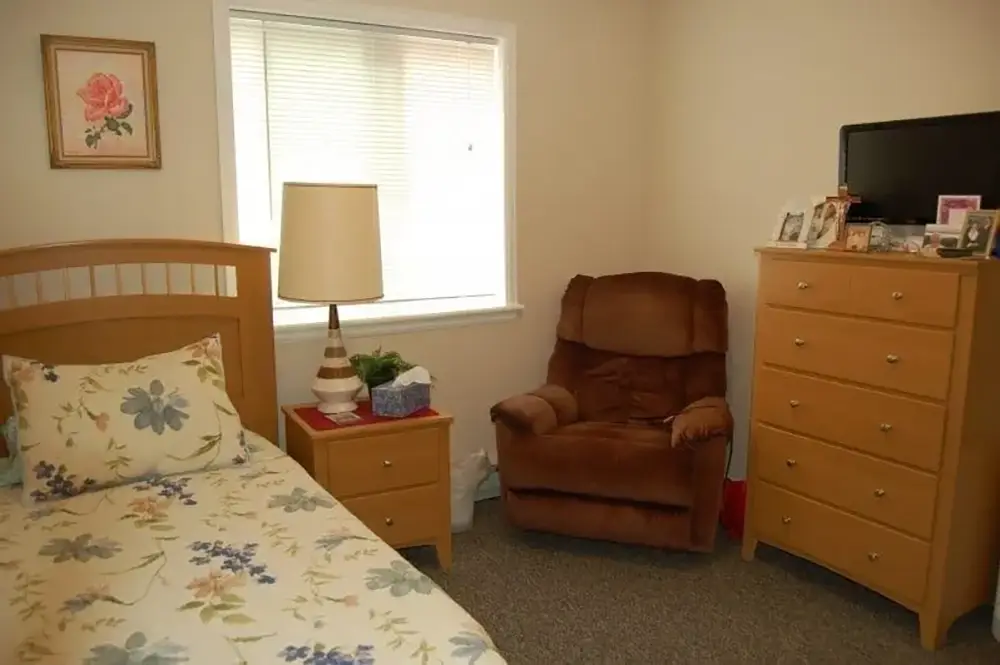 A bedroom with a single bed, floral bedding, wooden nightstand, lamp, brown recliner, tall dresser, small TV, and framed photos under a window with blinds.