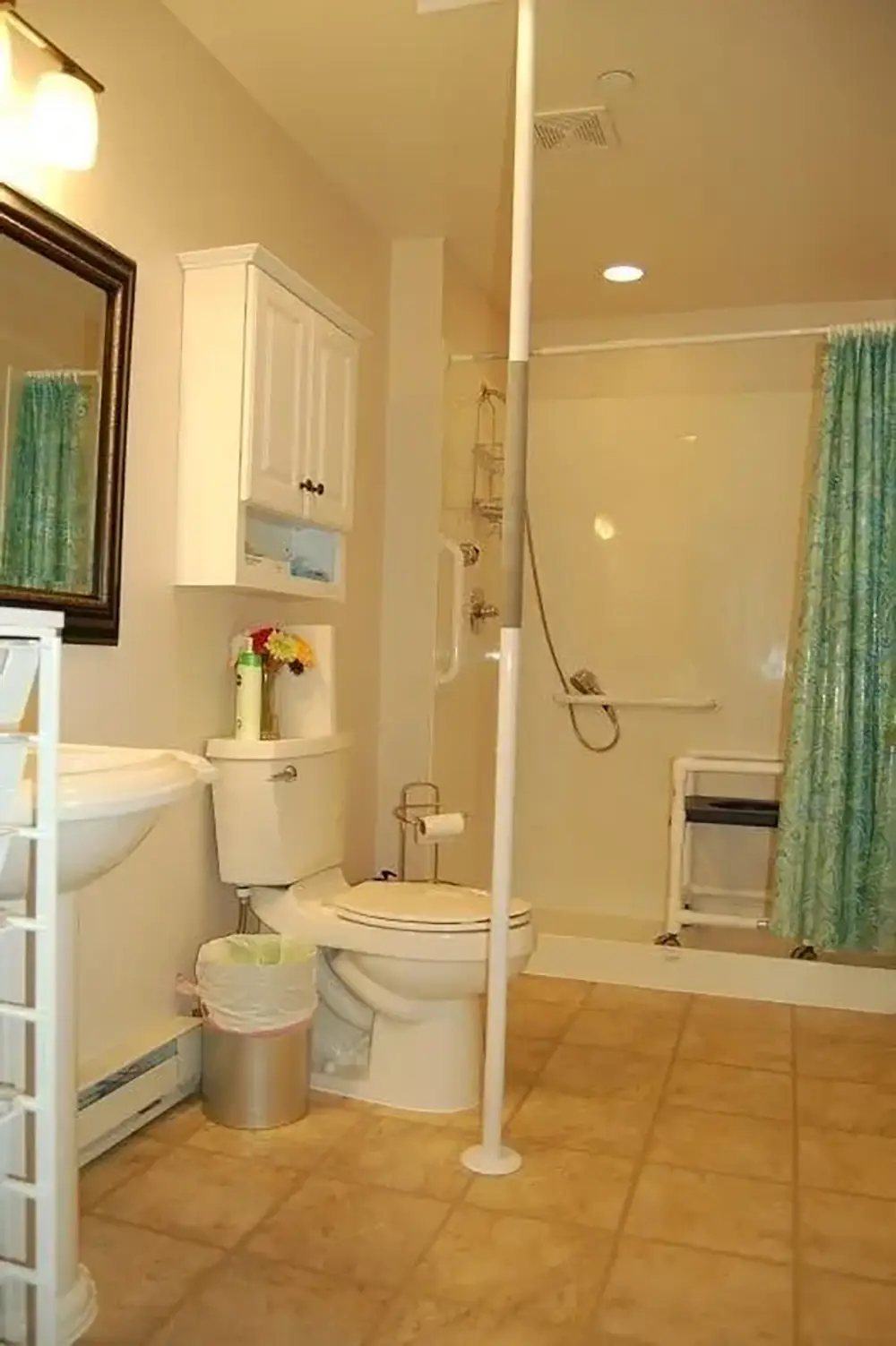 Bathroom with tiled floor, toilet, sink, wall cabinet, trash can, and walk-in shower with blue curtain and a shower seat. A vertical pole stands between the toilet and sink.