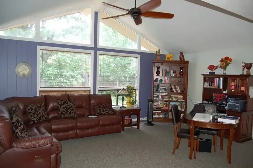 Living room with brown leather sectional, wooden bookcases, round dining table with chairs, ceiling fan, large windows, and blue accent wall.