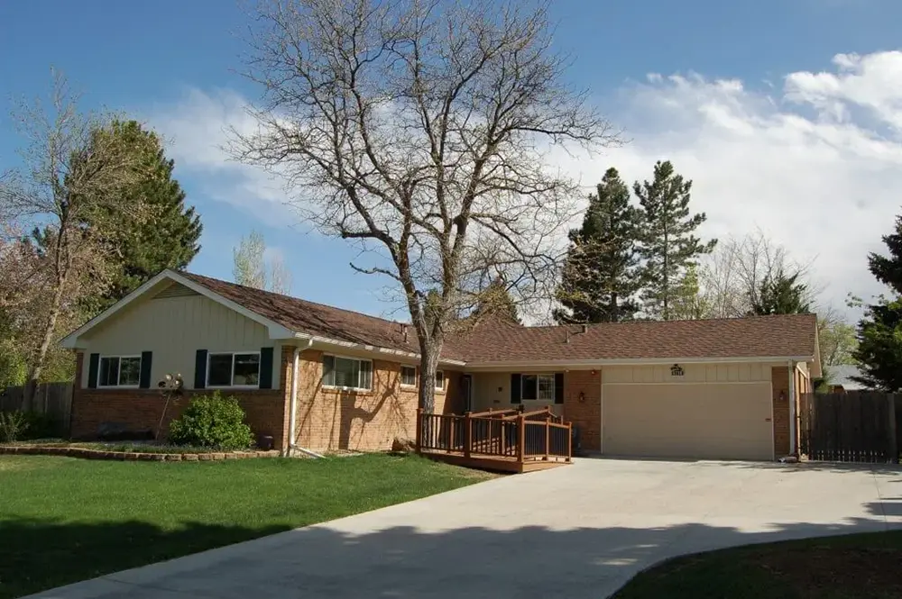 Single-story brick house with attached garage, ramp to the front door, a leafless tree in the front yard, green grass, and a concrete driveway under a partly cloudy sky.
