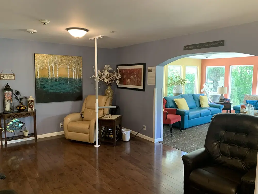 Living room with wood floors, a tan recliner, artwork, and a white support pole; archway leads to a sunroom with blue sofa, windows, and bright orange walls.