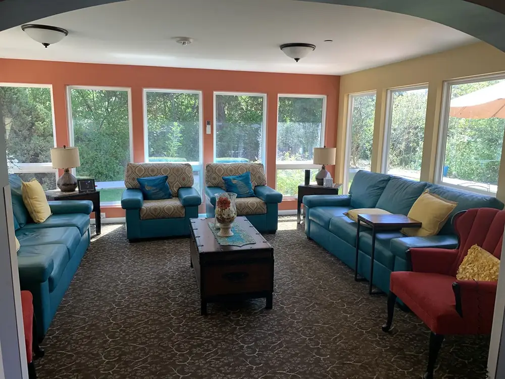 A sunroom with large windows, blue sofas, two armchairs, side tables with lamps, and a central coffee table on a patterned carpet.