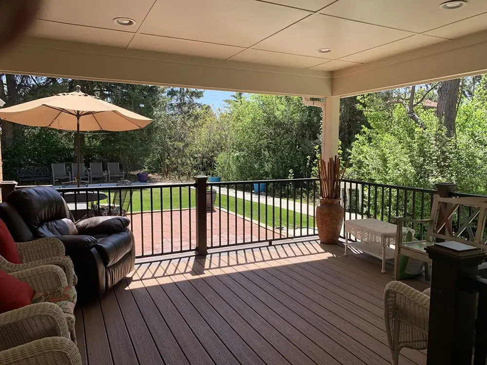 Covered patio with wicker and leather seating, overlooking a backyard with grass, trees, a large umbrella, and outdoor lounge chairs.