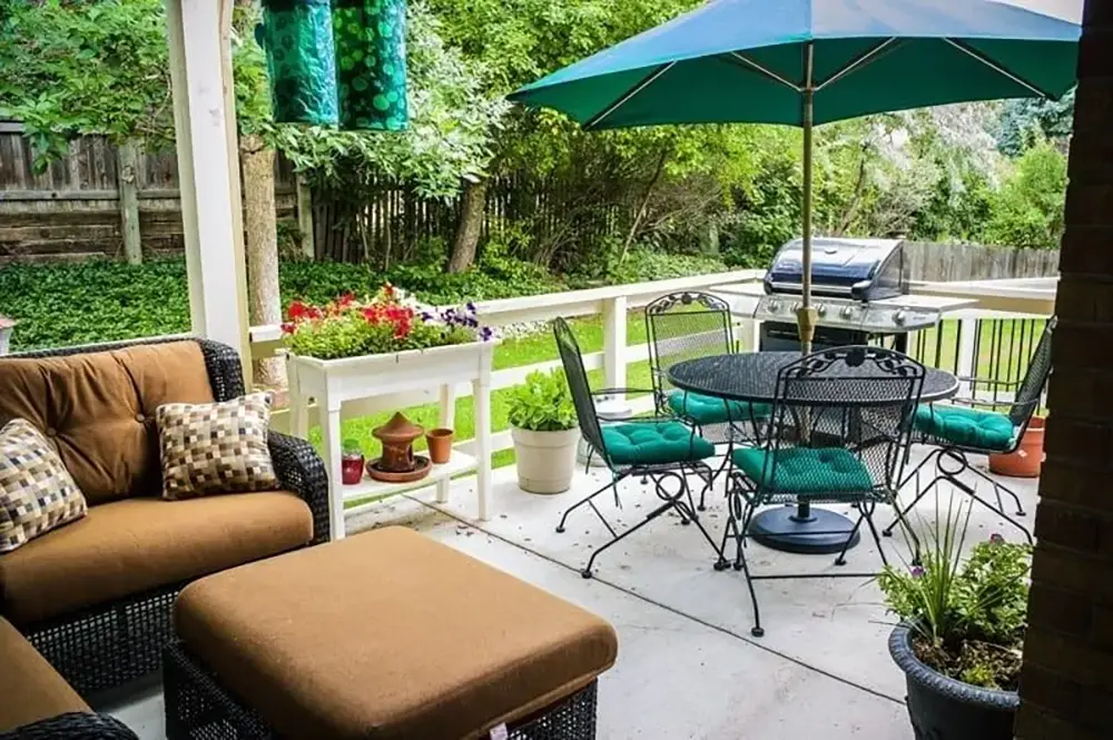 Patio with brown wicker furniture, a black metal table and chairs with green cushions, a large umbrella, potted plants, a barbecue grill, and a grassy yard in the background.