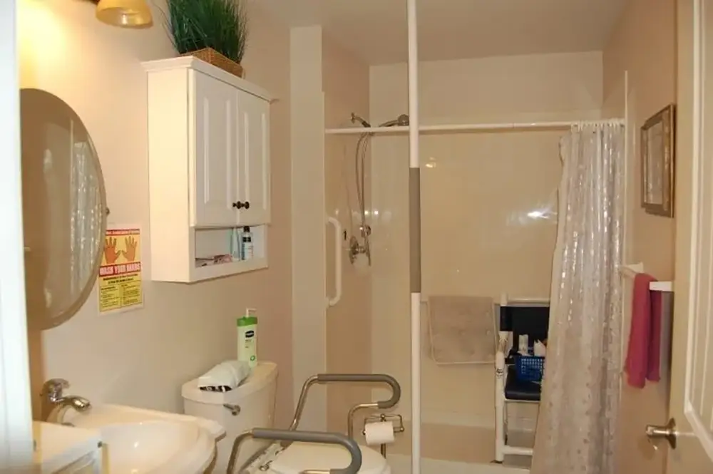 Accessible bathroom with grab bars, a walk-in shower with a chair, toilet with support rails, white cabinets, and a sink. Towel and toiletries are visible.