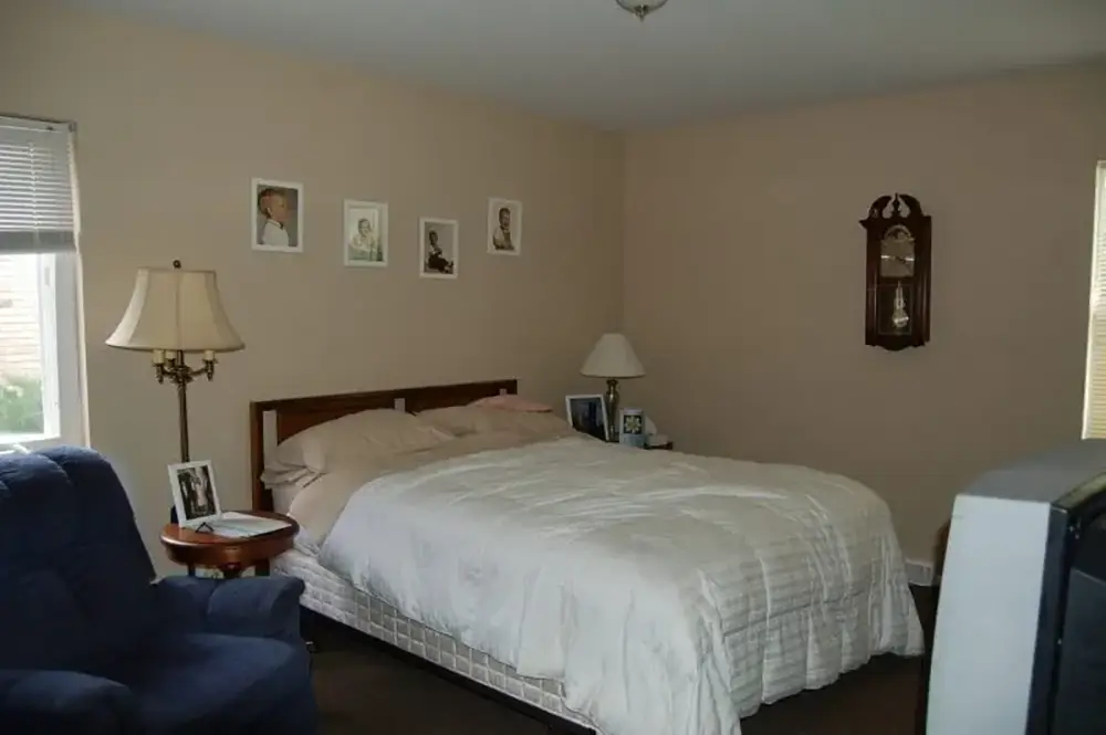 A neatly made bed in a simple bedroom with beige walls, two lamps, framed photos, a wall clock, a blue chair, and a window letting in natural light.