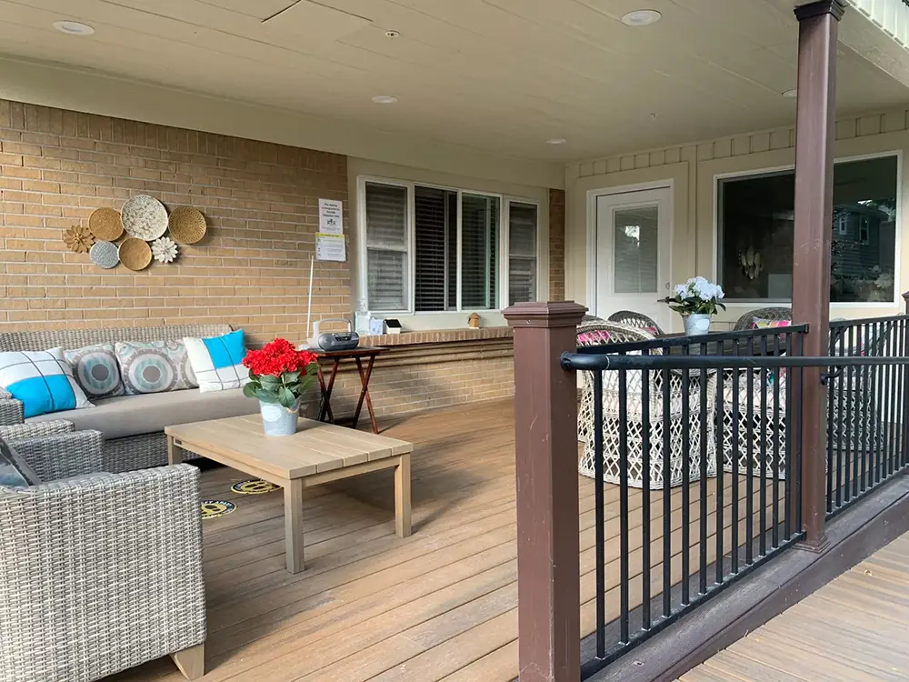 Covered porch with wicker furniture, colorful cushions, potted flowers on a wooden table, wall decor, and a black railing in front. Large windows and a door are visible on the brick wall.