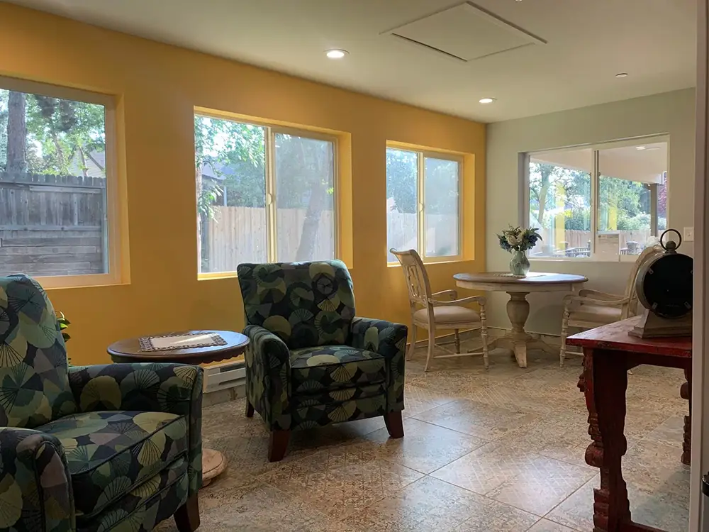 Sunny sitting room with two patterned armchairs, a small table with chairs, tiled floor, yellow walls, and large windows overlooking a fenced yard.