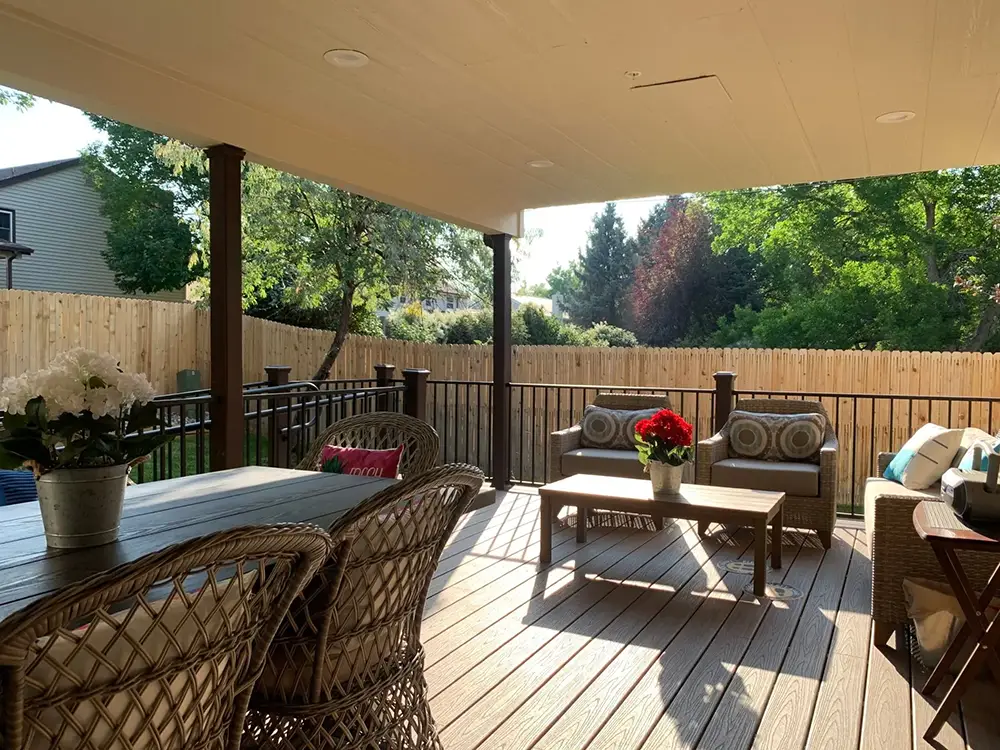 Covered backyard patio with wicker dining set, cushioned seating area, and potted flowers, enclosed by a wooden fence and surrounded by trees in daylight.