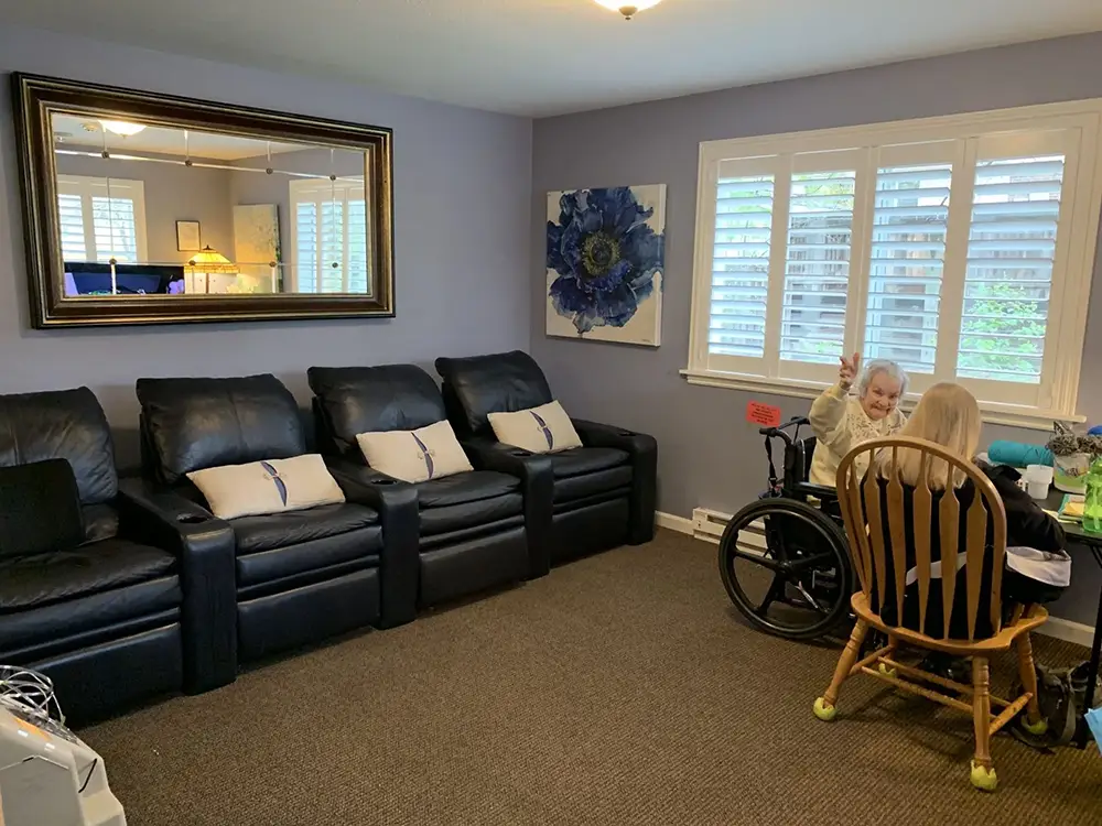 A woman in a wheelchair sits at a table with another person in a living room with black recliners, a large mirror, a flower painting, and white shuttered windows.