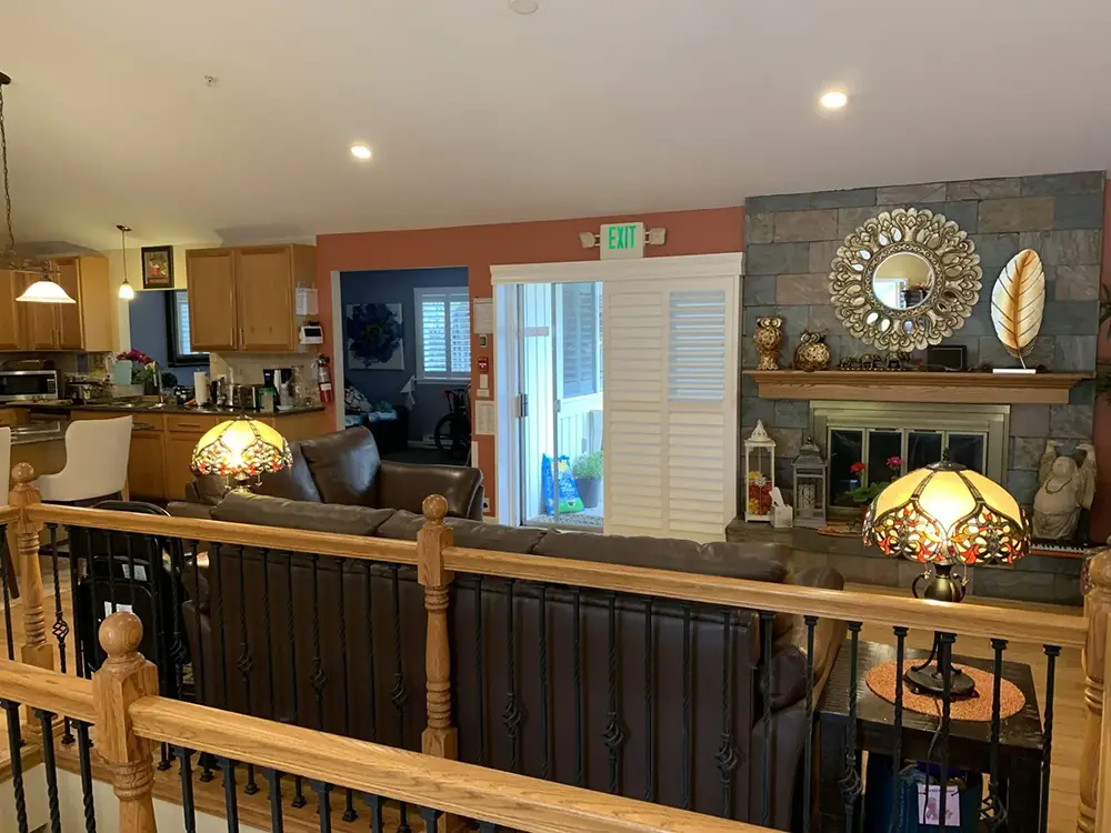 A living room with brown couches, a stone fireplace, decorative lamps, and a mirror above the mantel. The kitchen is visible to the left and a glass door leads to another room.