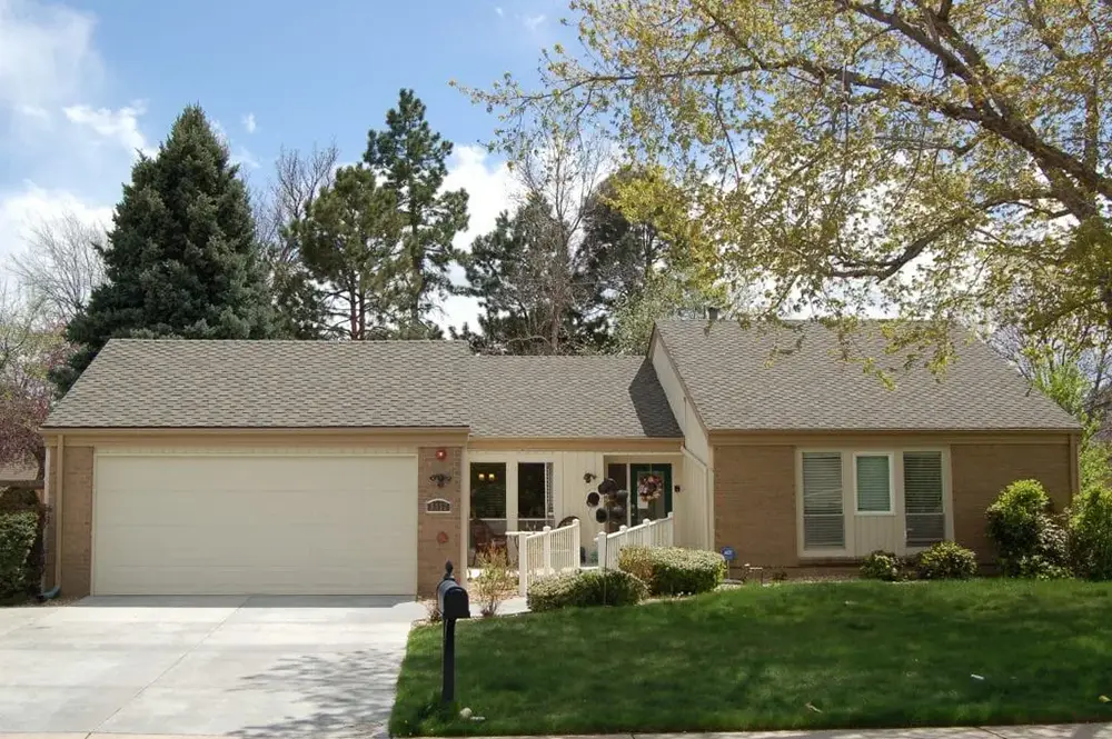 Single-story suburban house with attached two-car garage, light brown brick exterior, large front lawn, and tree partially shading the yard on a sunny day.