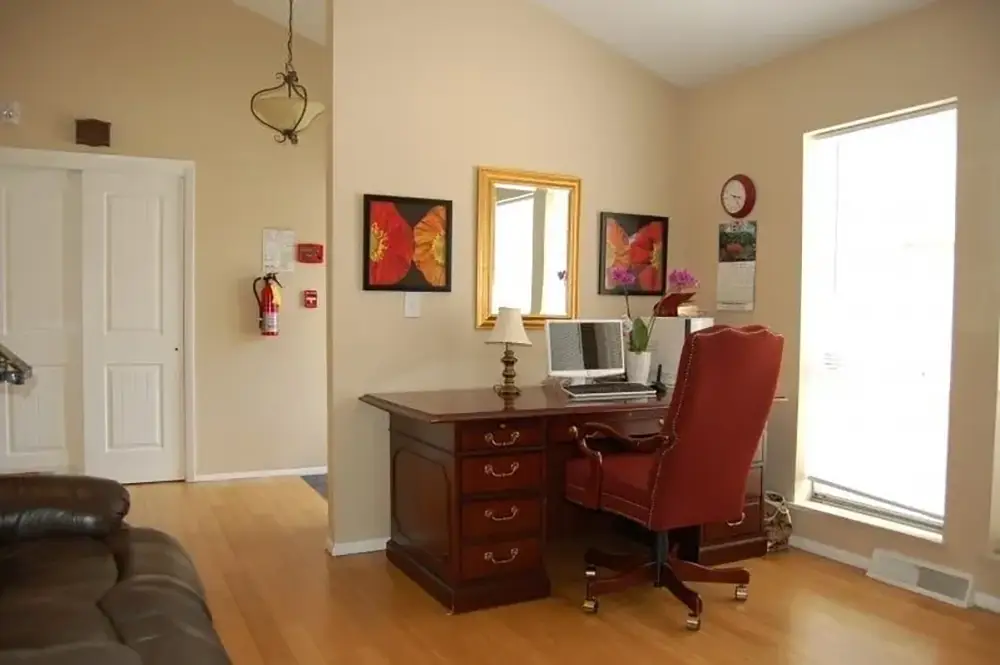 Home office with a wooden desk, red chair, desktop computer, lamp, and butterfly artwork. There is a large window, calendar, and wall clock nearby. Light wood floor and beige walls.