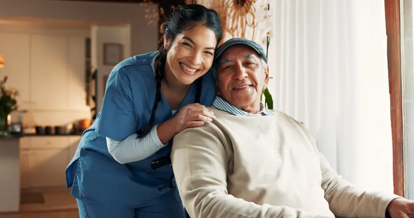 A nurse stands behind an older man sitting in a chair by a window, both smiling at the camera in a well-lit home setting.