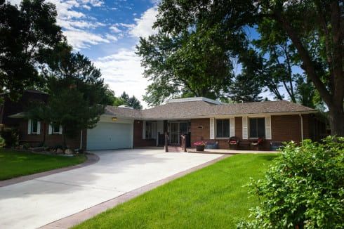 Single-story brick house with attached garage, large driveway, manicured lawn, and mature trees under a partly cloudy sky—an inviting alternative to large assisted living facilities.