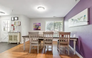 Dining area with a wooden table, four chairs, light hardwood floor, purple accent wall, bird artwork, window with trees outside, and entryway visible to the left.