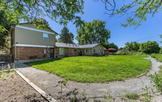Two-story brick and siding house with large backyard, circular lawn, outdoor seating area, and surrounding trees under a clear blue sky. Curved walking path borders the grassy area.