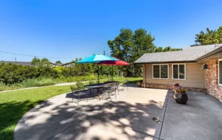 Concrete patio with a table, chairs, and a large multicolored umbrella next to a house, surrounded by grass and trees under a clear blue sky.