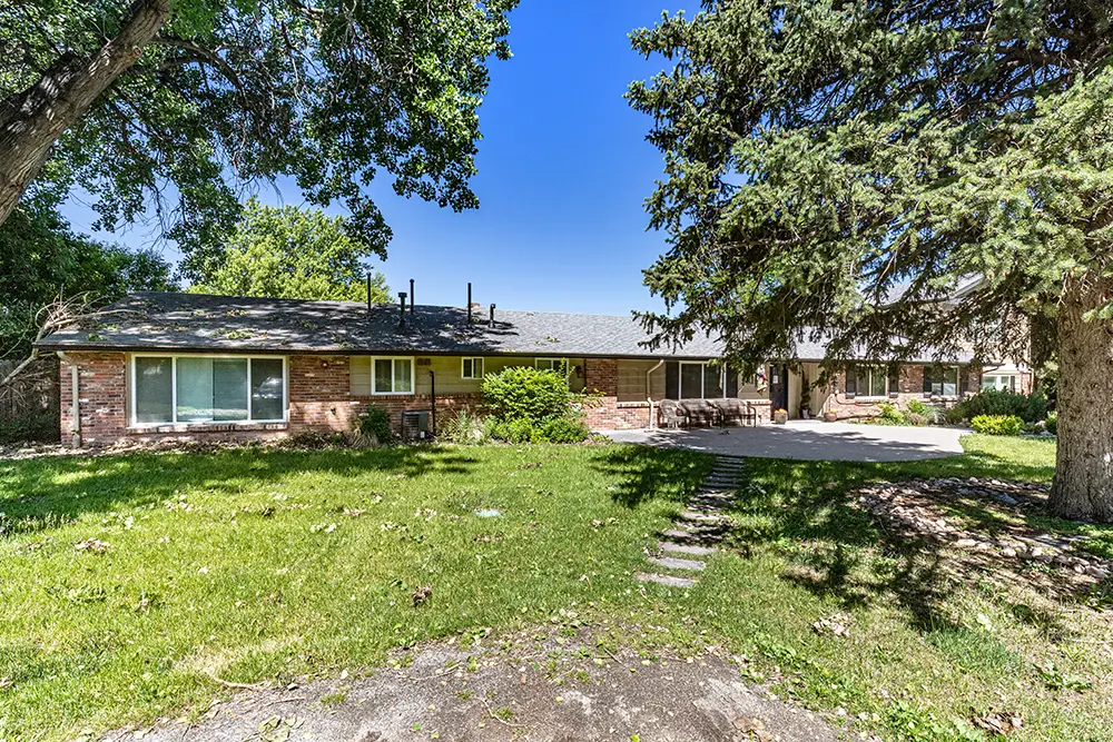 Single-story brick house with large windows and a gray shingle roof, surrounded by a grassy yard and mature trees under a clear blue sky.