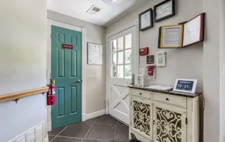 Small entryway with a teal door labeled "Activities," a white cabinet, wall-mounted documents, clock showing Monday 10:31, fire extinguisher, and glass door leading outside.