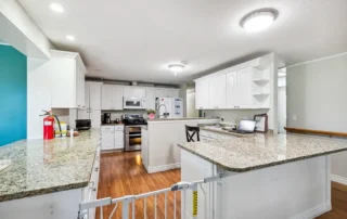 Modern kitchen with white cabinets, granite countertops, stainless steel appliances, wood flooring, and a laptop on the counter. A baby safety gate is installed at the foreground.