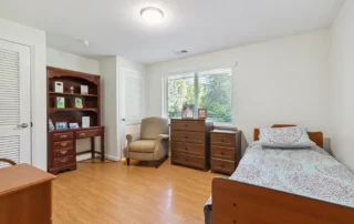 A tidy bedroom with a single bed, wooden dressers, bookshelf, armchair, desk, and a window letting in natural light.