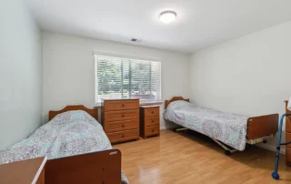 Bedroom with two single beds, three dressers, light wood floor, window with blinds, and white walls; beds have matching patterned bedspreads.