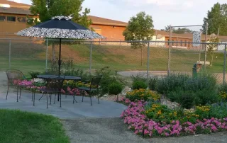 Littleton-gallery-10 A patio set with four chairs and an umbrella is placed on a concrete pad next to a flower garden, enclosed by a chain-link fence near school buildings.