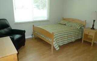 Littleton-gallery-2 A simple bedroom with a wooden bed, striped bedding, a nightstand with a lamp, a desk, and a black armchair on a light wood floor.