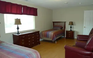 Littleton-gallery-4 A bedroom with two single beds, two red armchairs, a dresser with two lamps, and a window with a red valance.