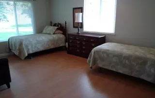 Littleton-gallery-6 A simple bedroom with two single beds, a dark wooden dresser with a lamp and mirror, large windows, and a brown armchair on a wood floor.
