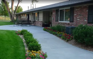 Littleton-gallery-7 A wide concrete walkway leads to a single-story brick and siding building, bordered by green lawn, shrubs, and colorful flowerbeds. Outdoor benches and tables sit near the entrance.