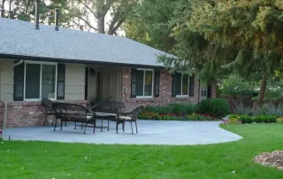 Littleton-gallery-9 A backyard patio with a metal table and four chairs on a concrete surface, adjacent to a brick house with large windows and surrounded by green grass and landscaping.