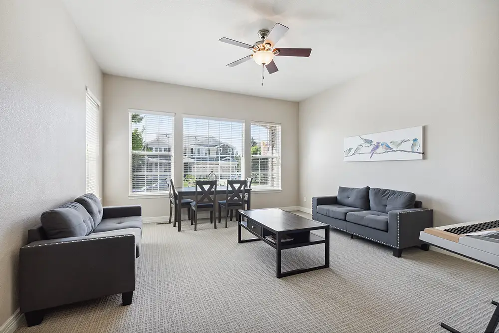 Bright living room with two gray sofas, a coffee table, a dining table with four chairs, wall art, and large windows with blinds. Ceiling fan overhead.