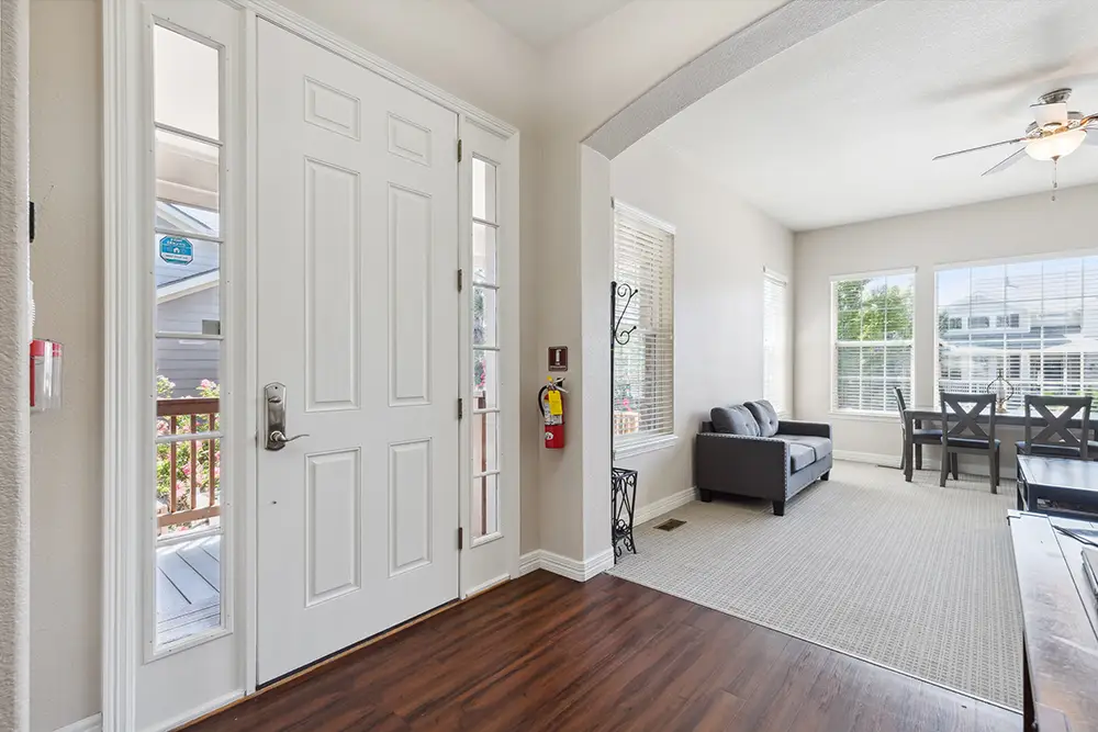Bright entryway leads to a living and dining area with large windows, a ceiling fan, a gray sofa, a dining table with chairs, and hardwood flooring transitioning to carpet.
