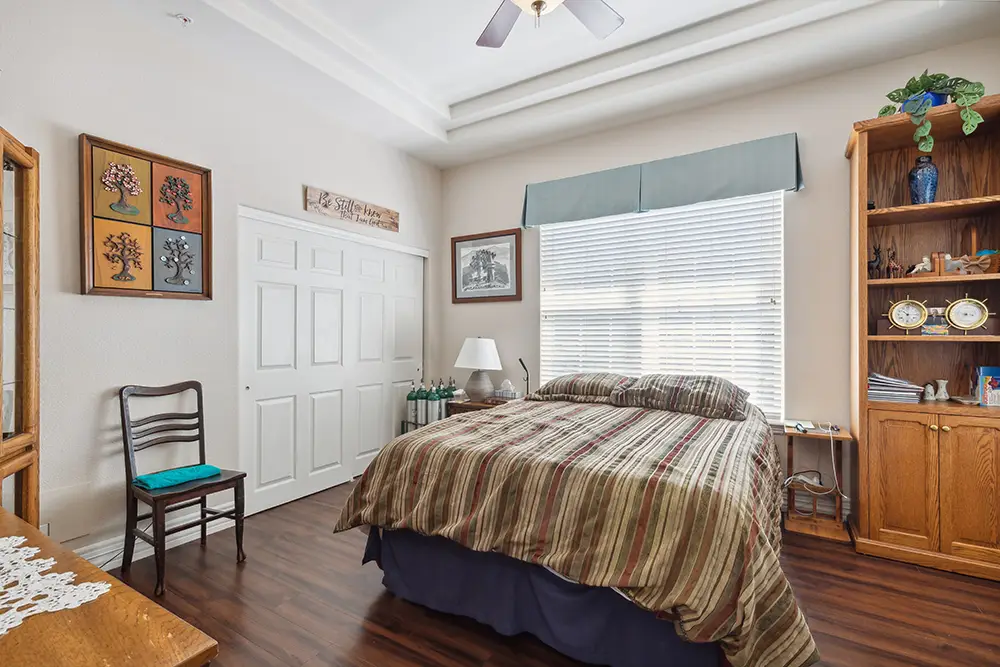 Bedroom with a striped bedspread, wooden furniture, wall art, a window with blinds, and a ceiling fan overhead. The room has wood flooring and decorative items on the shelves.