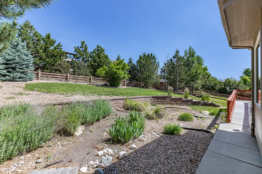 A backyard with gravel landscaping, sparse plants, stone pathway, sloped grass area, retaining wall, wooden fence, and trees under a clear blue sky.
