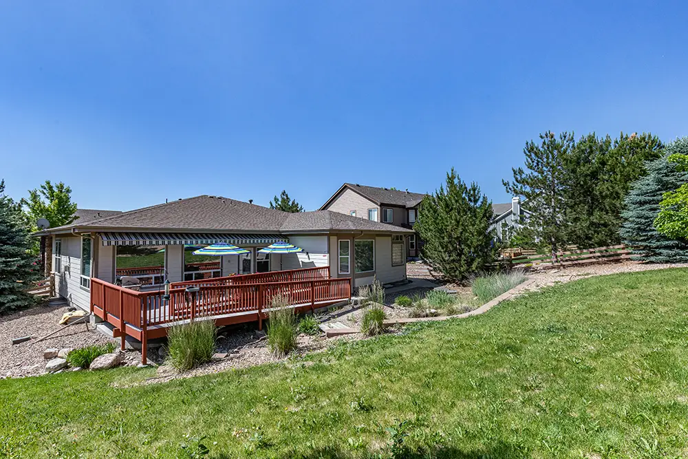Single-story house with a red deck, striped umbrellas, and a spacious backyard featuring grass, shrubs, and trees under a clear blue sky.