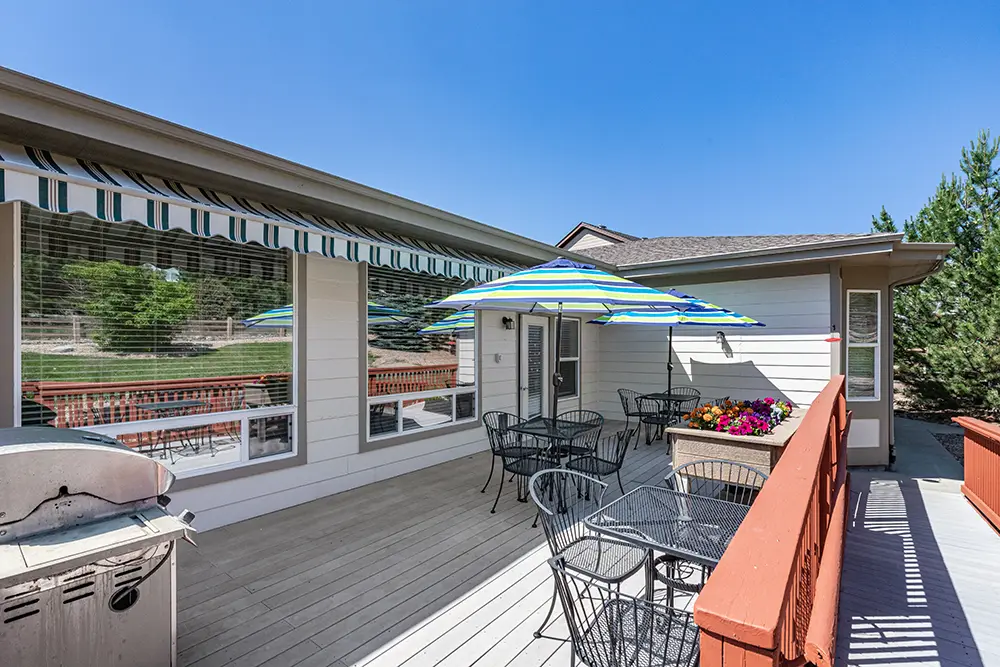 Outdoor patio with metal tables and chairs, striped umbrellas, a planter with flowers, a barbecue grill, and large windows reflecting greenery under a clear blue sky.